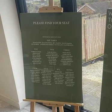Two green signs on easels with wedding details, one showing seating chart and the other wedding information, in front of a window.